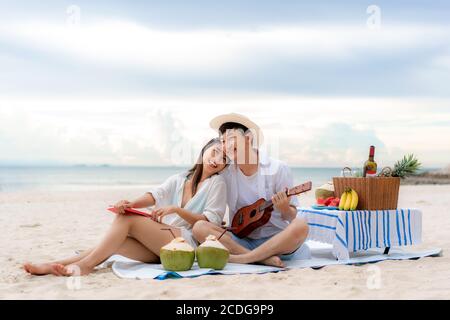 Asian couple happy and enjoy the trip honeymoon of couple lover on the sea beach by playing Ukulele and singing song music together. Summer, holidays, Stock Photo
