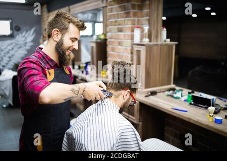 Young bearded man getting haircut by hairdresser with electric razor at barber shop Stock Photo