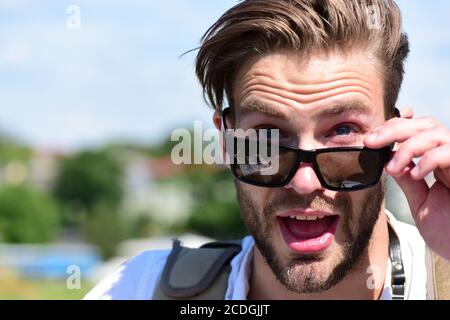 Man with beard and open surprised face wears sunglasses. Tourist on blurred cityscape background, copy space. Traveller looks for tourists attractions. Travelling and vacations concept. Stock Photo