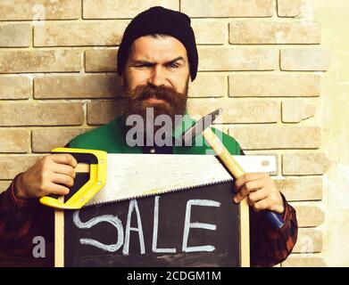 bearded foreman, long beard, brutal caucasian hipster with moustache holding various building tools: saw, hammer and board with inscription sale, happy smiling face, brick wall studio background Stock Photo