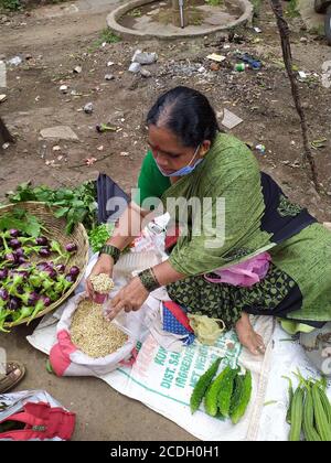 Aged lady wear face mask and selling vegetables in market yard. Measuring moth bean legumes with use of measure utensil.. Stock Photo