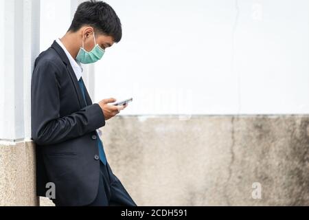 A handsome young asian business man using smartphone. Asian young attractive businessman wearing a face mask leaning against the wall. Stock Photo