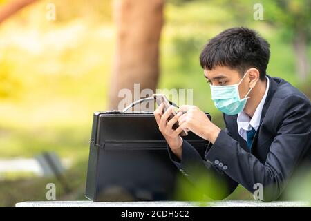 A handsome young asian business man using smartphone. Asian young attractive businessman wearing a face mask sitting on the table. Stock Photo