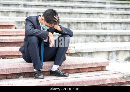 Failure unemployed stressed young asian business man in suit and wearing a face mask covering head with hands. A young businessman sitting desperate o Stock Photo