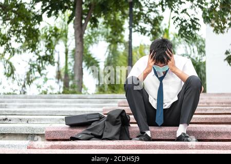 Failure unemployed stressed young asian business man wearing a face mask covering head with hands. A young businessman sitting desperate on the stairs Stock Photo