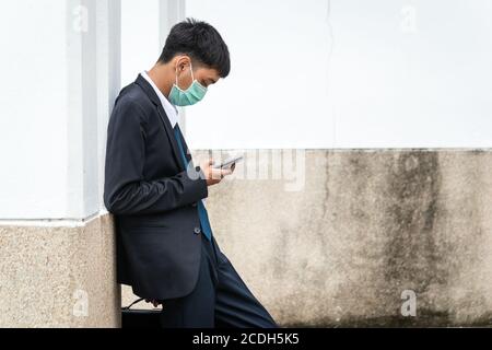 A handsome young asian business man using smartphone. Asian young attractive businessman wearing a face mask leaning against the wall. Stock Photo