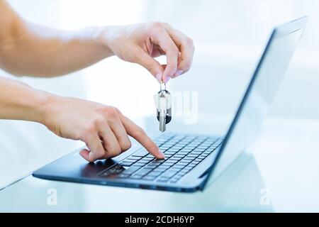 Close-up of male person's hands holding up keys while typing on a laptop computer keyboard. Online booking and rentals concept. Stock Photo