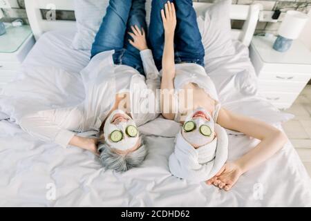 Top angle view of two charming women, senior gray haired lady and young girl with towel on head, lying on the bed and enjoying spa day with face masks Stock Photo