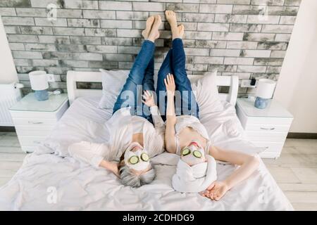 Top angle view of two charming women, senior gray haired lady and young girl with towel on head, lying on the bed and enjoying spa day with face masks Stock Photo