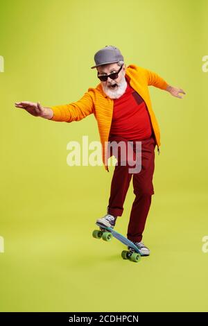 Moving skate. Portrait of senior hipster man in fashionable eyewear isolated on green studio background. Tech and joyful elderly lifestyle concept. Trendy colors, forever youth. Copyspace for your ad. Stock Photo
