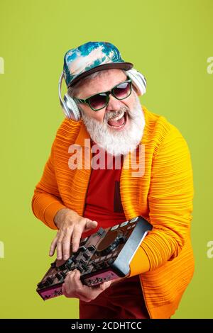 Party guy. Portrait of senior hipster man in fashionable eyewear isolated on green studio background. Tech and joyful elderly lifestyle concept. Trendy colors, forever youth. Copyspace for your ad.