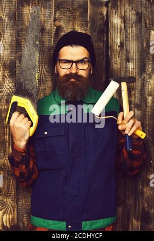 bearded painter, long beard, brutal caucasian hipster with moustache holding various building tools: saw, hammer, roller paint with serious face on brown wooden studio background Stock Photo