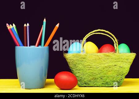Eggs and pencils. Coloured crayons in blue mug and Easter decorations in green basket and red egg lying on yellow wooden table isolated on black background. Childhood and Easter concept Stock Photo