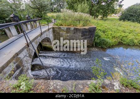 Keutelbeek stream with a small bridge over the dam and sluice with its black metal railing surrounded by green vegetation in the Sittard city park Stock Photo