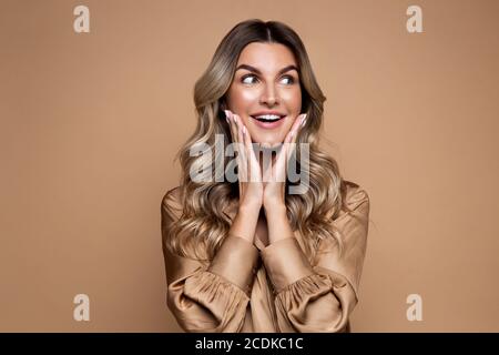 Surprised blonde woman with curly hair dressed in silk shirt, smiles pleasantly, looking aside at blank space for your advertisement, happy with sale Stock Photo