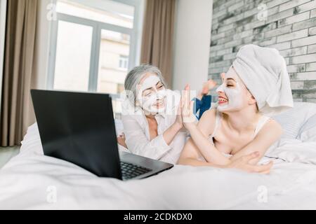 Two women with facial masks, pretty young girl wrapped in towel and her senior attractive grandmother, having fun while watching movie on laptop at Stock Photo