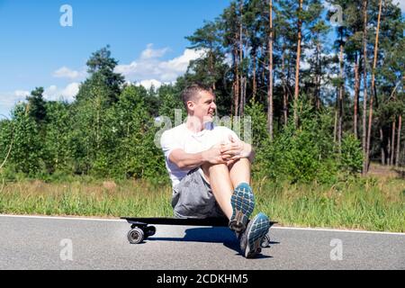 Young adult man enjoys the weather sitting on a skateboard against the backdrop of the forest Stock Photo