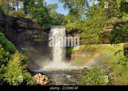 A majestic waterfall framed by lush foliage on a sunny day. A rainbow is visible in the mist near the base of the falls. Stock Photo