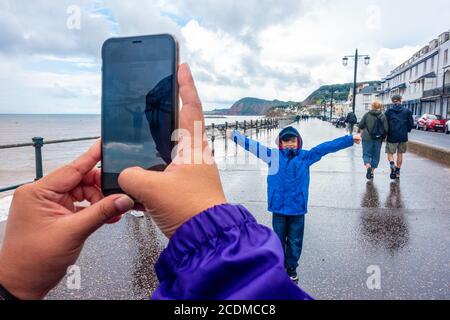 Close up of a lady taking a photo of her son using a mobile phone on the esplanade at the seafront at Sidmouth in South Devon, UK on a wet day. Stock Photo