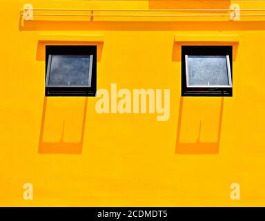 A plain bright yellow plastered wall with two open windows and casting shadows from the overhead midday sun. Stock Photo