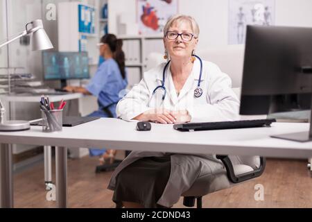 Mature medical therapist sitting at desk in hospital cabinet while nurse in blue uniform working on computer. Stock Photo