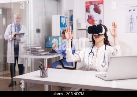 Young doctor using augmented reality goggles in hospital office while senior medic is taking notes on clipboard in clinic corridor and nurse working on computer. Stock Photo