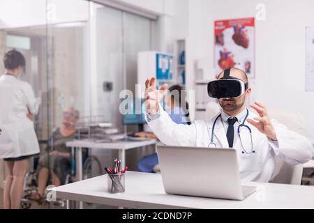 Doctor using futuristic augmented reality technology wearing headset in hospital office and white coat with stethoscope. Disabled senior woman discussing with medic on clinic corridor. Stock Photo