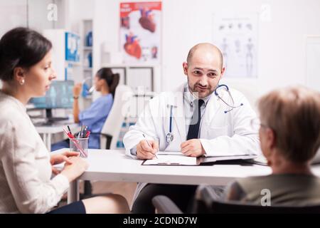 Mature woman with walking disablity in wheelchair with daughter during medical check with doctor in hospital office. Medic writing notes on clipboard and nurse is working on computer. Stock Photo