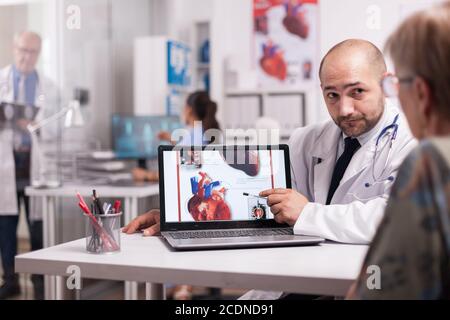 Doctor pointing at heart with trauma after senior patient heart attack. Elderly aged woman discussing about medication with young medic in hospital office. Mature medic wearing white coat and taking notes on clipboard. Stock Photo