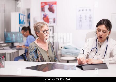 Doctor using tablet computer while examining senior woman in hospital office. Medic discussing diagnosis with patient about her illness. Stock Photo