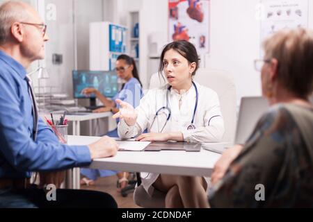 Female doctor explaining diagnosis to senior man elderly woman in hospital office during medical check up. Sick married pensioners. Nurse working on computer. Stock Photo