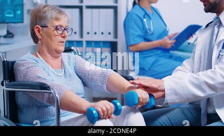 Physiotherapy for elderly woman in wheelchair, she is practicing with dumbbells to recover after a musle trauma. Modern private hospital or rehabilitation clinic. Lifting exercise for disabled people Stock Photo