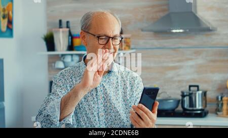 Seniore retired man saying hello during a video call using smarhphone in kitchen while having breakfast. Elderly person using internet online chat technology video webcam making a video call connection camera communication conference call Stock Photo