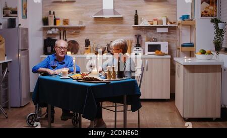 Happy senior elderly wife eating romantic dinner at home in the cozy kitchen with her disabled husband. Imobilized paralyzed handicapped elderly man having romantic dinner. Stock Photo