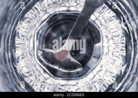 Metal teaspoon in a faceted glass cup with reflection, top view, close-up. Gray and blue abstract background, futuristic wallpaper, transparent surfac Stock Photo