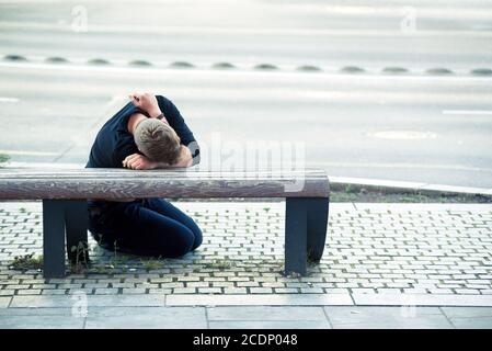 Broken man sitting on a ground resting his head on a bench Stock Photo