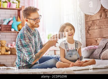 Happy loving family. Father is combing her daughter's hair sitting on the bed in the room. Stock Photo
