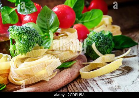 Raw pasta fettuccine, fresh broccoli, green basil leaves, red cherry tomatoes, vintage wooden background, selective focus Stock Photo