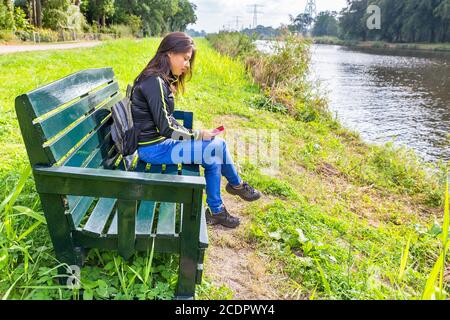 Woman operating mobile phone on bench at river Stock Photo