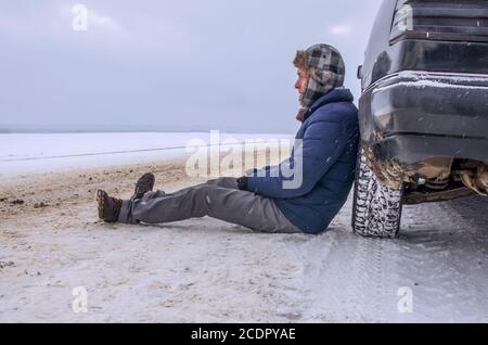 Angry young man waiting for help, sitting near the broken car on the side of the road in the winter in the woods Stock Photo