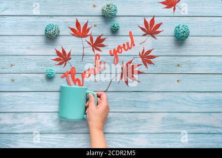 Hand holds ceramic mug with words Thank you cut out of paper. Seasonal Fall flat lay with Autumn decorations on light grey aged wood, maple leaves and Stock Photo