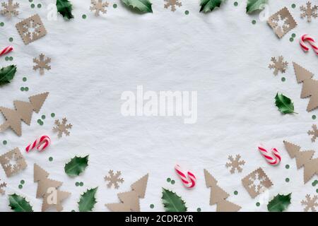 Christmas flat lay, top view on white textile. Eco friendly decor from craft paper, candy canes and natural holly leaves. Xmas tree silhouettes, snowf Stock Photo