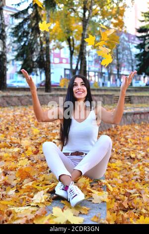 Beautiful girl in autumn park sitting on a concrete parapet catches falling maple leaves with her hands, a smile on her face, in white jeans and a blo Stock Photo