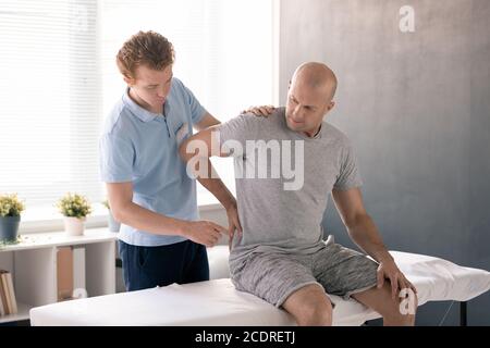 Bald mature man touching painful back while sitting on medical couch in clinics Stock Photo