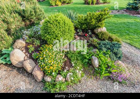 Landscaped summer garden with green plants, rocks, flowers in flowerbeds, mown grass. Stock Photo