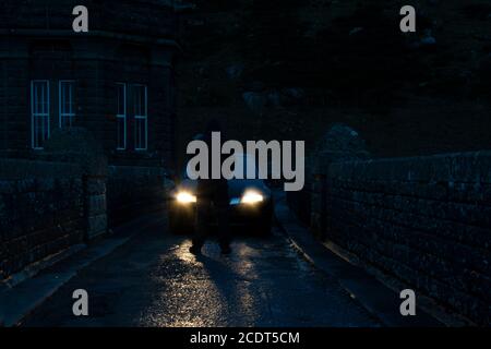 A mysterious hooded man, back to camera. Standing in front of car headlights. On a country lane at night Stock Photo