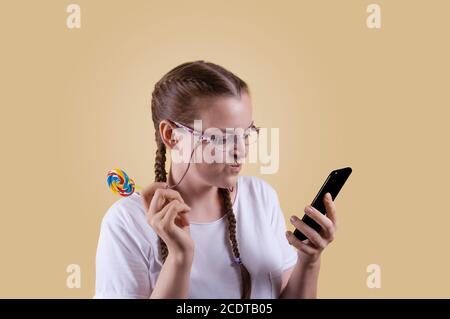 The girl looks at the phone. The girl is holding the phone. Girl with pigtails. The girl is shot against a yellow background. The girl smiles at the n Stock Photo