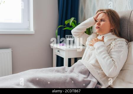 Beautiful blonde young woman have strong headache and chest pain. Female sits in bed and hold head with her hands, suffering on her face Stock Photo