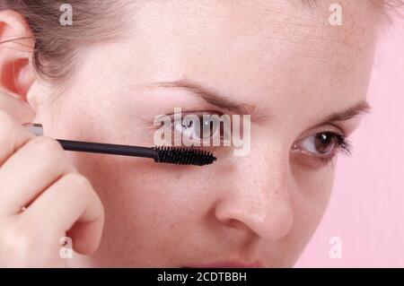 The girl looks in the mirror and holds an eyelash brush. Girl paints eyelashes. A nice girl looks in a palette mirror and paints eyelashes. Big brown Stock Photo