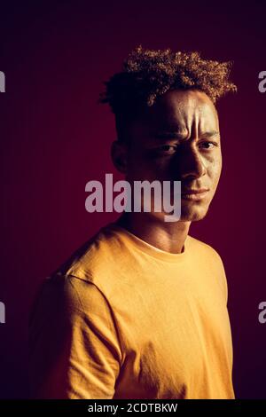 Freestyle. Mulatto guy standing isolated on dark red in yellow light looking camera doubtful close-up Stock Photo